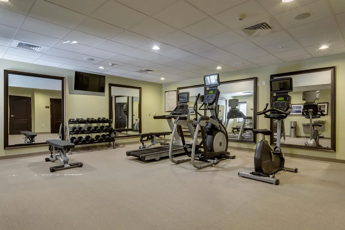 A bright, energizing fitness center featuring an elliptical machine, exercise bike, dumbbell rack, weight bench, and wall mirrors. The atmosphere is motivating, with ample equipment for various workout routines.