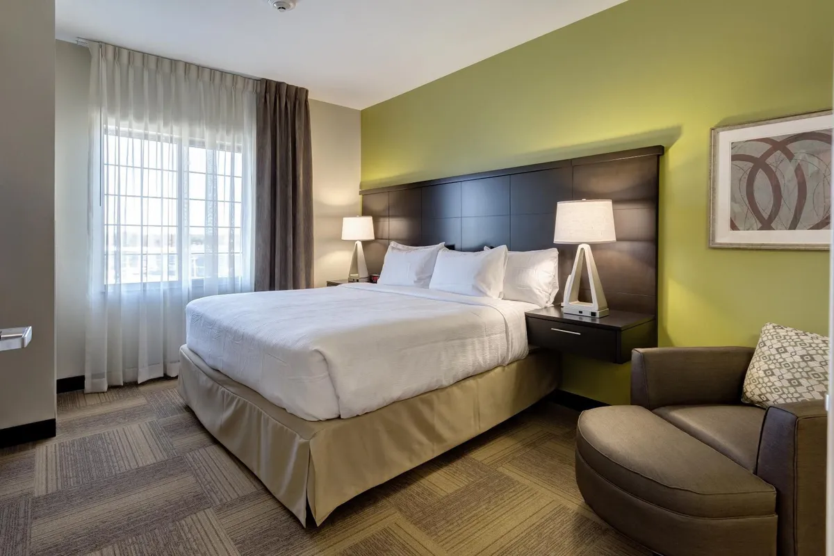A cozy king bedroom featuring a plush bed, an inviting armchair next to a large window, and a softly glowing lamp. The room is bathed in natural light, creating a peaceful and restful ambiance ideal for unwinding after a busy day.