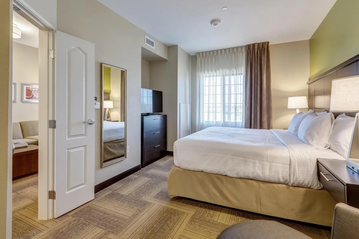 A well-appointed suite that combines a comfortable bed and a living area with a sofa, a television, and a window letting in bright sunlight. The space is decorated in a modern style, offering areas for both relaxation and entertainment.