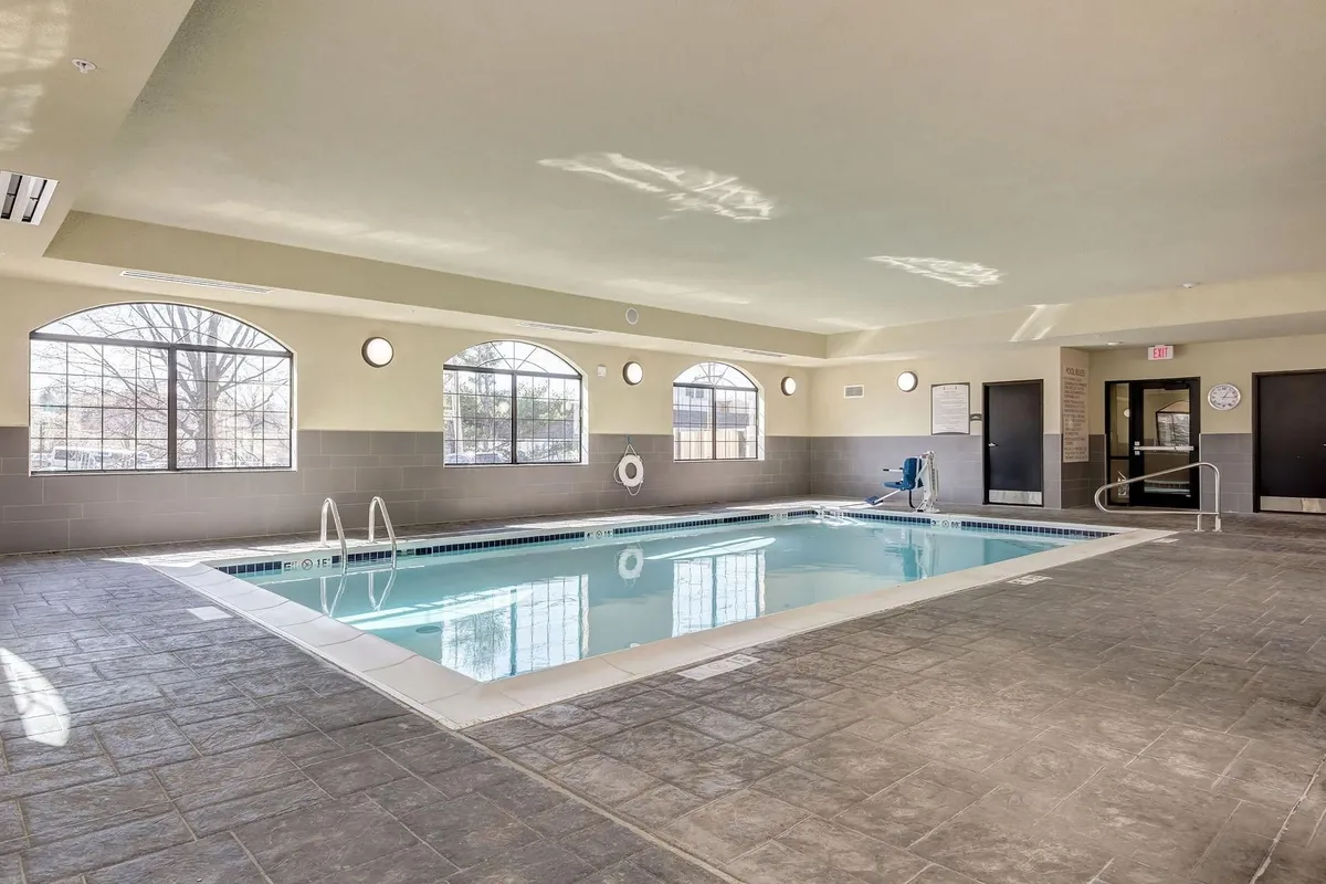 This image showcases a bright and inviting swimming pool area with large windows letting in natural light. Safety equipment is visible for peace of mind, and the setting suggests a space for relaxation or gentle exercise. The pool is clean, with comfortable seating areas and a calming ambiance, perfect for guests needing restful moments away from the hospital environment.